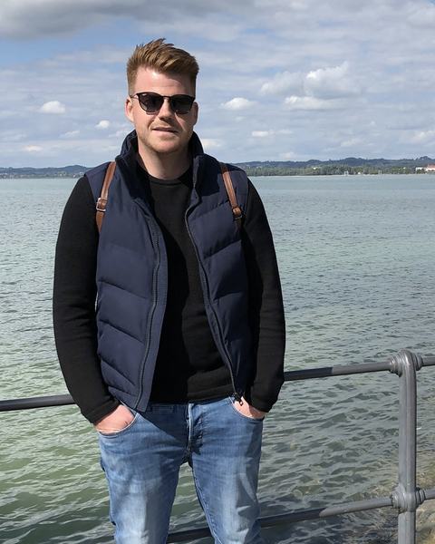 Ben standing on a lakeside promenade, wearing sunglasses and a dark vest, with the lake and a cloudy sky behind him.