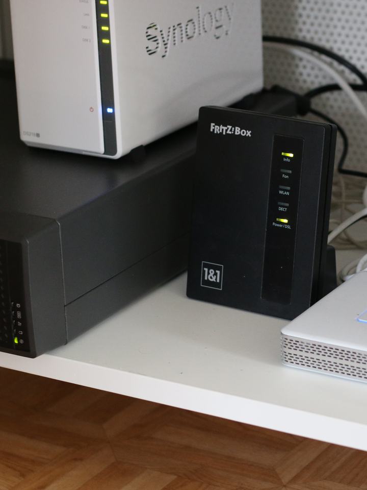 A white two-bay Synology NAS on top of a black Fujitsu TX120 S3 tower server, next to a Fritz!Box router and a Raspberry Pi in a tangle of white cables.