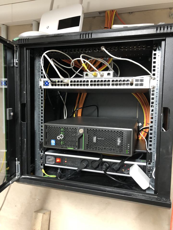 A small black wall cabinet with a network switch, a patch panel and a single Fujitsu server.