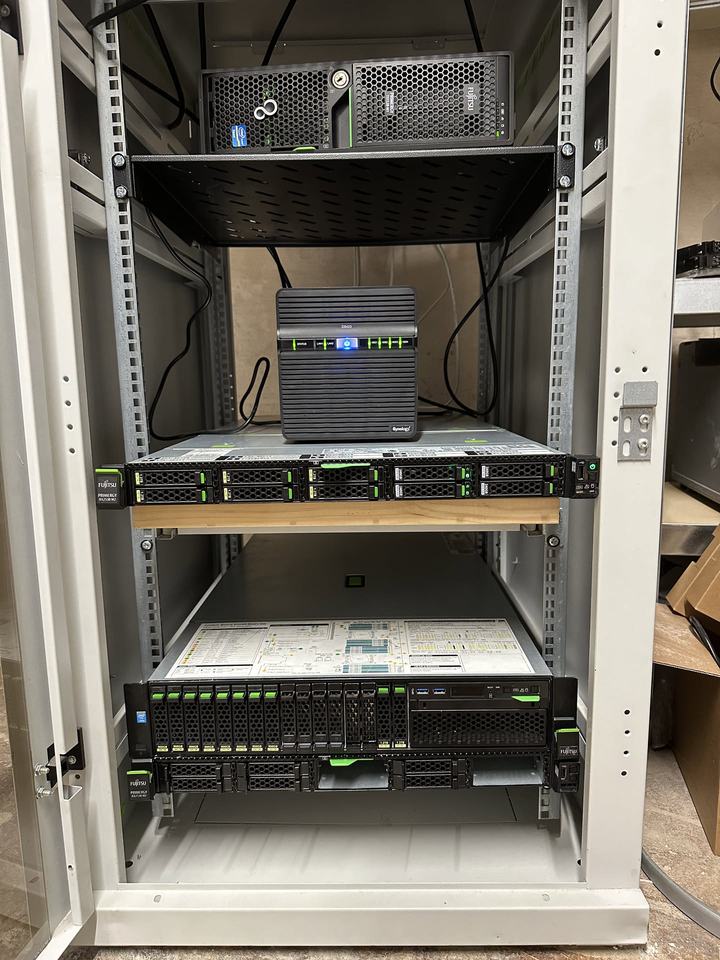 A grey 19-inch rack with a Synology NAS on a shelf above two Fujitsu rack servers.