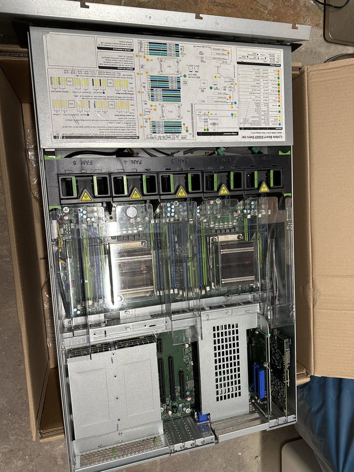 The opened Fujitsu RX2540 M1 rack server in its cardboard box, with two heat sinks, memory slots, fans and the PCIe riser cards visible.