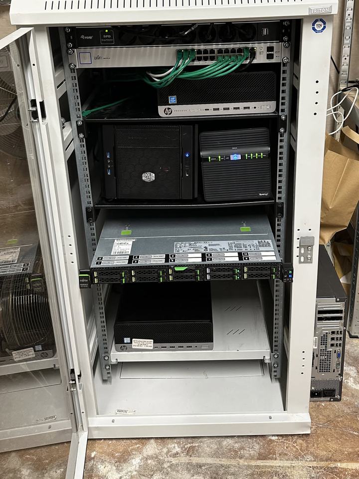 A grey rack cabinet with a switch, small PCs, a Synology NAS and a rack server, the glass door open.